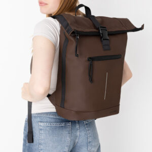 A person with long hair wears a white shirt and blue jeans, carrying a brown New York Rolltop Rucksack - Mocha featuring a front zip pocket and black buckle straps, standing against a plain white background. Gate21.ee