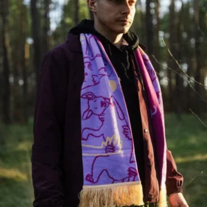 A person stands in a sunlit forest wearing the Puppy - Organic scarf, which features colourful pink drawings and a yellow fringe. They also wear a green cap, burgundy jacket, and dark hoodie, with trees and grass visible in the background. Gate21.ee