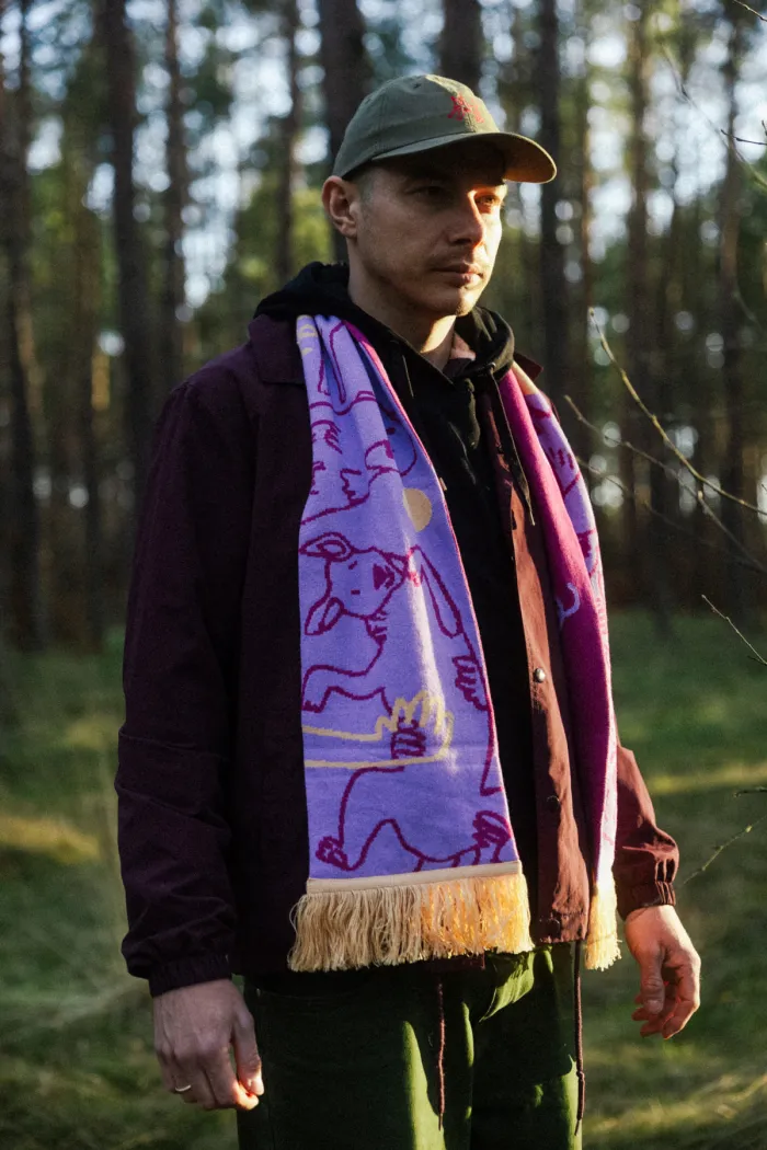 A person stands in a sunlit forest wearing the Puppy - Organic scarf, which features colourful pink drawings and a yellow fringe. They also wear a green cap, burgundy jacket, and dark hoodie, with trees and grass visible in the background. Gate21.ee