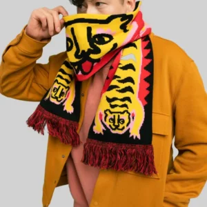 A person wearing a mustard jacket and pink shirt covers their lower face with the “Puppy - Organic” scarf, which features a yellow, black, and red design with tassels. The background is plain light grey. Gate21.ee