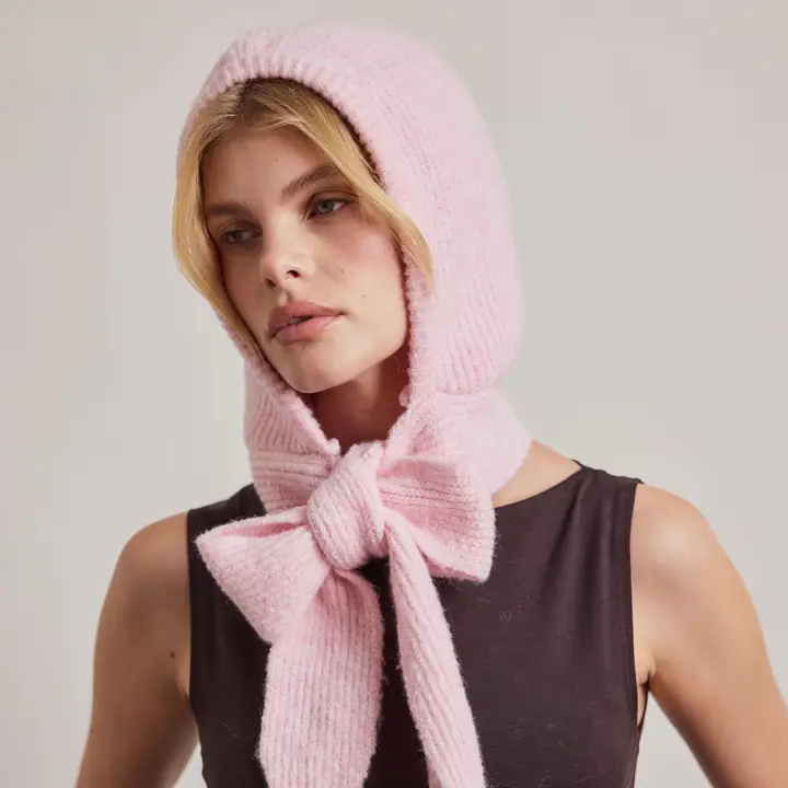 Wearing a sleeveless black top and The Bonnet - Light Pink - Recycled—a textured pink hood with a large bow tied under the chin—complemented by cosy Arctic Fox Reykjavik hand warmers, the person gazes off to the side against a plain background. Gate21.ee