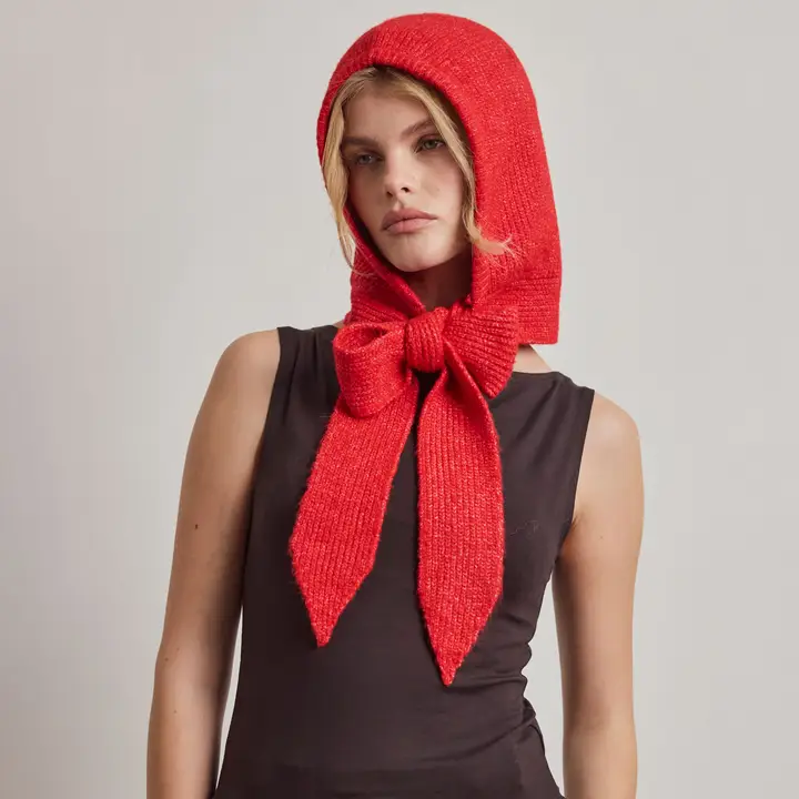 Wearing The Bonnet - Red - Recycled, a person stands against a plain background, the vibrant recycled knit hood tied in a large bow under the chin and thoughtfully styled with cosy Arctic Fox Reykjavik hand warmers and a sleeveless black top for added warmth and flair. Gate21.ee