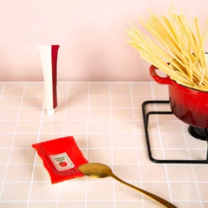 A red “Ketchup - Plate” pot holding uncooked spaghetti sits on a stand, evoking the artistry of L’expressionist gifts. Nearby, a gold spoon, a small red packet labelled tomato, and a capped tube are neatly arranged on a tiled counter against a pale pink wall in the background. Gate21.ee