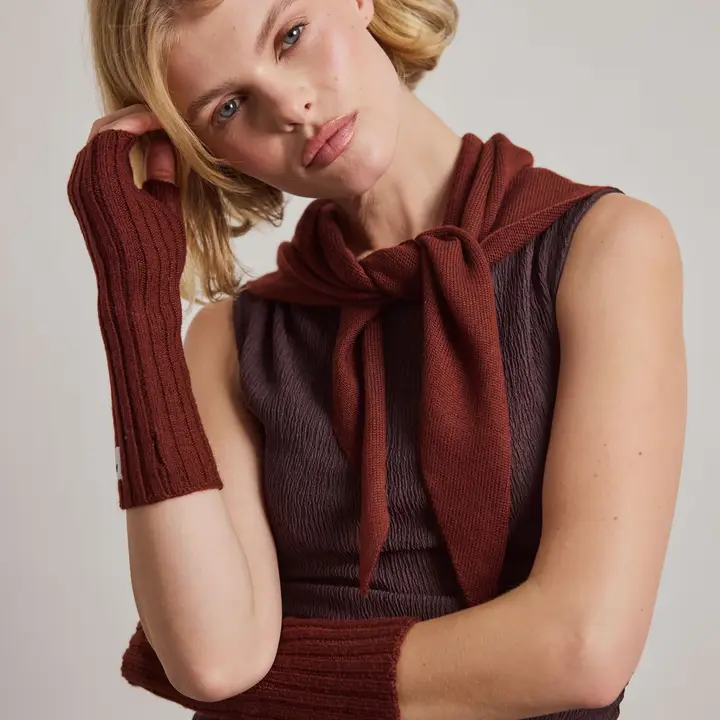 A woman with short blonde hair wears a sleeveless textured top, a rust-coloured jumper tied around her shoulders, and matching Merino Wool Fingerless Gloves in dark walnut brown. She rests her hand on the side of her head and looks at the camera. Gate21.ee