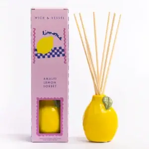 A lemon-shaped ceramic diffuser with reed sticks stands next to a pink and purple Happy Cat - 100ml Diffuser box labelled Wick & Vessel. This elegant Wick & Vessel home fragrance set features a charming box adorned with a lemon illustration and a window that reveals the beautifully crafted Happy Cat diffuser inside. Gate21.ee
